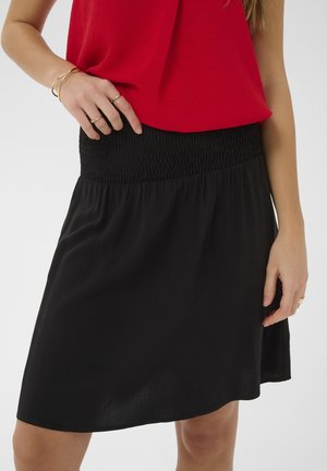 Vrouw draagt een rood mouwloos topje en een zwarte rok met geplooide taille, toont gouden ringen en armbanden aan haar handen.