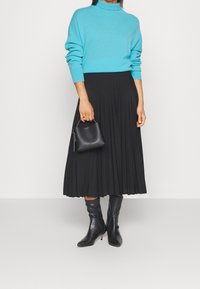 Pull turquoise à col montant, associé à une jupe midi plissée noire. Le mannequin tient un petit sac à main noir. Bottes noires montantes.