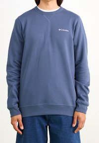 Szaro-niebieski sweatshirt wykonany z miękkiego materiału, z okrągłym dekoltem, ściągaczami przy rękawach oraz małym białym logo Columbii na piersi.