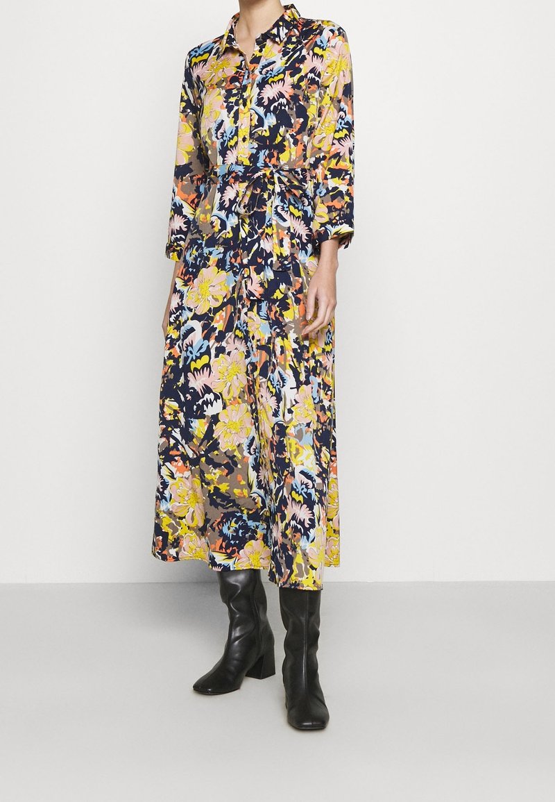 Femme portant une robe fleurie à manches longues, longue jusqu’aux chevilles, avec une ceinture à la taille et des bottes noires à talons montant jusqu’aux genoux, debout devant un fond uni.