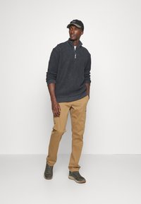 Dunkelgrauer Reißverschluss-Sweatshirt, khakifarbene Cargo-Hose, grüne Schuhe mit strukturierter Oberfläche, schwarze Kappe mit Logo; lässige Passform, lässiger Stil.