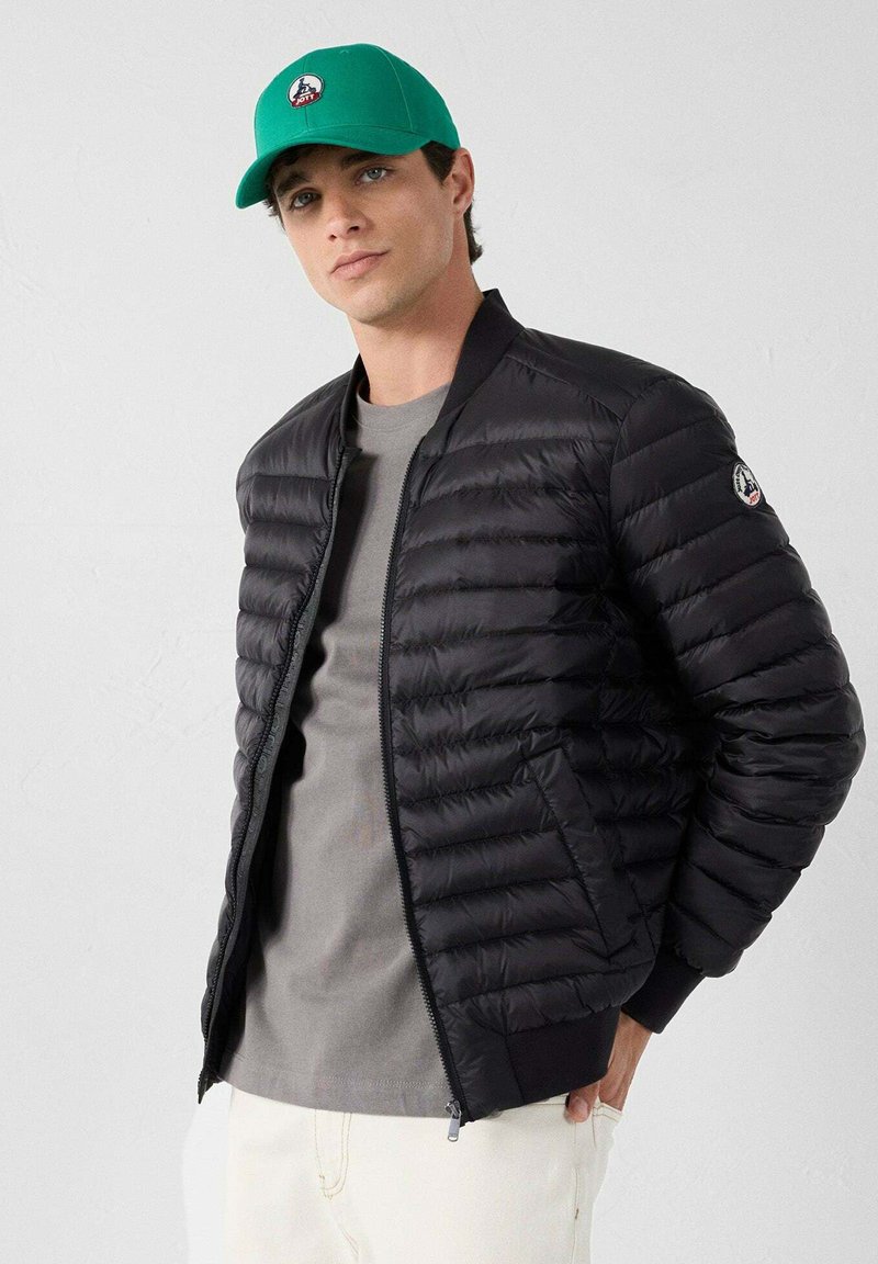 Chaqueta acolchada negra con diseño acolchado vertical, cremallera frontal y puños acanalados. Se lleva sobre una camiseta gris, combinada con una gorra verde con un logotipo.