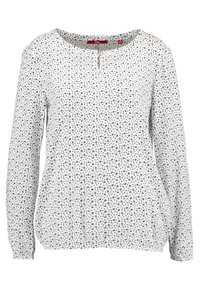 Haut blanc à manches longues avec un col rond, présentant un motif de petits points noirs et un détail de bouton au niveau du col. Tissu doux.