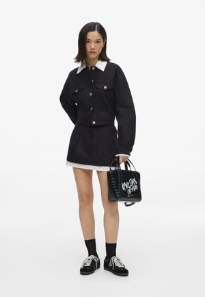 Joven mujer con chaqueta y falda negra con cuello y puños blancos, sosteniendo un bolso negro con texto blanco, de pie frente a un fondo blanco.