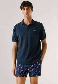 Noor mees, kes kannab tumesinist polosärki ja mereväesiniseid ujumispükse, millel on värvikad purjelaevade mustrid, seisab ühevärvilise heleda tausta ees.