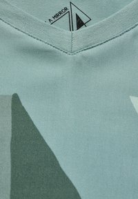 Jasnoniebieska koszulka z dekoltem w serek w geometryczne wzory w ciemnozielonym kolorze. Gładki materiał, wyraźne przeszycia i nadrukowana metka wewnątrz kołnierza.