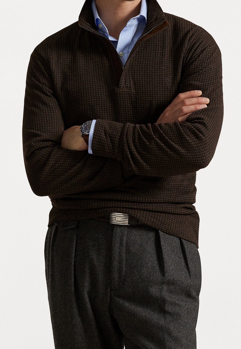 Homme portant un pull texturé marron foncé sur une chemise bleue à col, un pantalon plissé gris, une montre et une ceinture noire avec une boucle argentée.