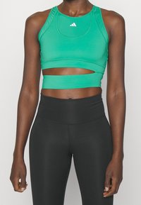 Zelený sportovní crop top s vysokým výstřihem, výřezem pod prsy a hladkým, elastickým materiálem. V kombinaci s černými legínami s vysokým pasem.