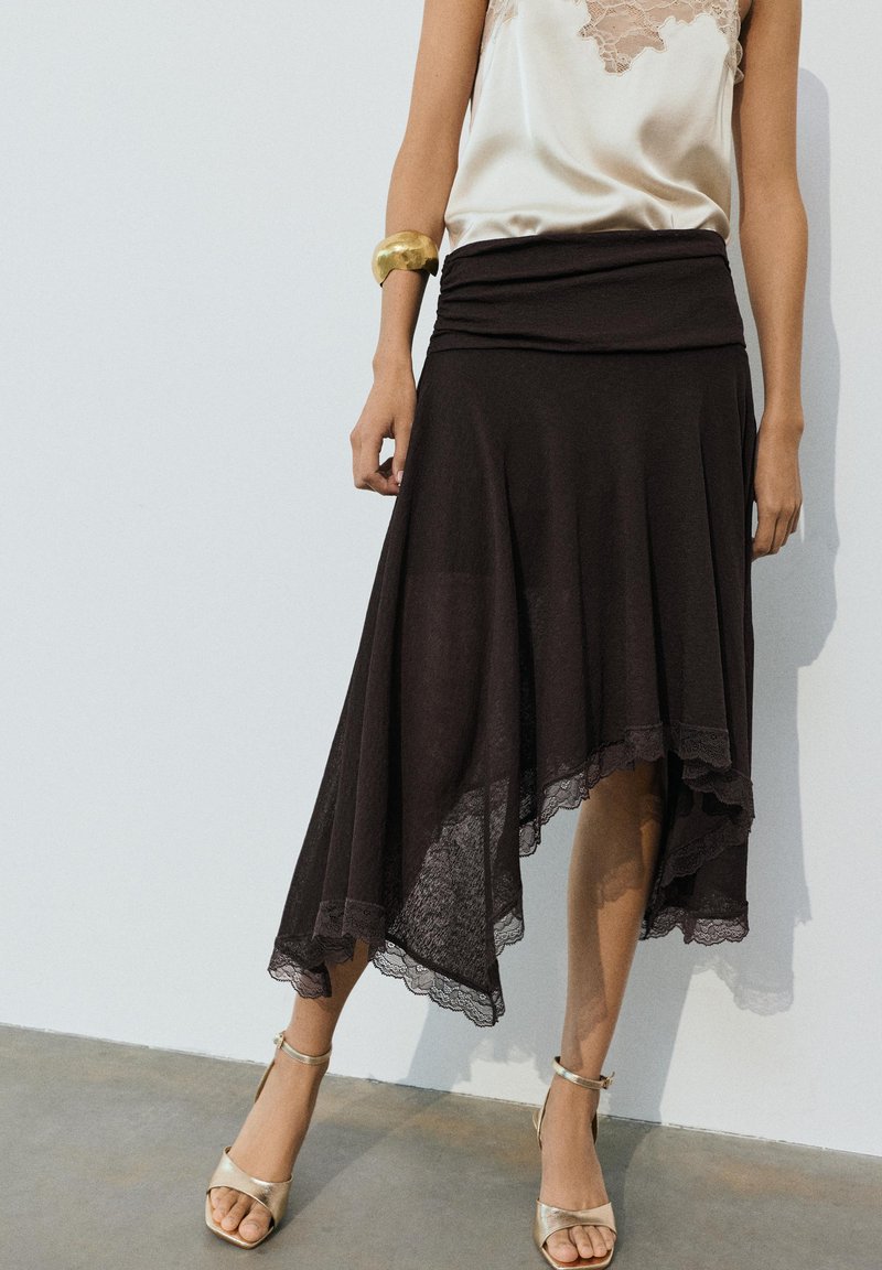 Femme portant une jupe asymétrique noire avec bordure en dentelle, un top en satin crème bordé de dentelle, un bracelet doré et des talons ouverts dorés, debout contre un mur blanc.