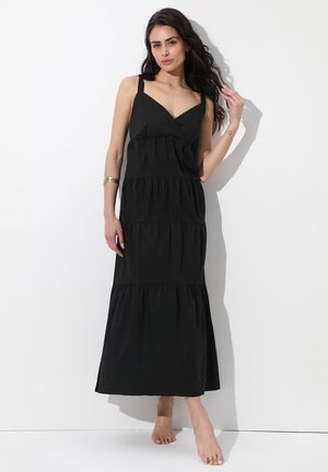 Donna con lunghi capelli scuri che indossa un abito nero lungo a più strati senza maniche, in piedi a piedi nudi contro un muro bianco con una mano che tocca i suoi capelli.