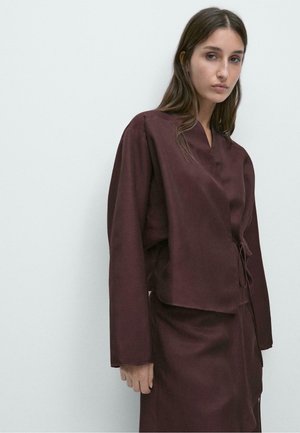 Mladá žena s dlouhými hnědými vlasy, oblečená v volném tmavě hnědém zavinovacím topu a odpovídající sukni, na jednobarevném bílém pozadí.