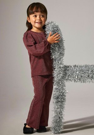 Jong meisje in een sprankelende maroon outfit dat glimlacht terwijl ze zilveren folie vasthoudt die om een stoel gewikkeld is, tegen een effen achtergrond.