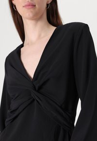 Čierne šaty s hlbokým výstrihom do V, prekrúteným predným dizajnom a dlhými rukávmi z hladkej, ľahkej látky. Minimalistický a elegantný vzhľad.