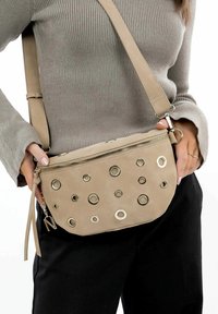 Beige crossbodytas met een gladde textuur, voorzien van ronde metalen accenten en een ritssluiting. Verstelbare riem en decoratieve tassel.
