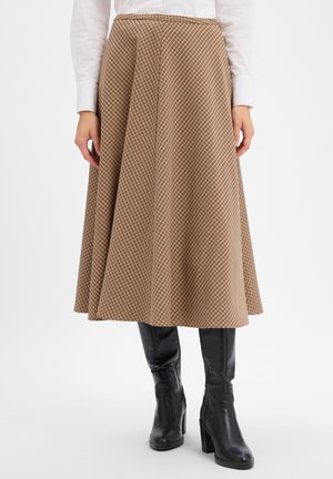 Vrouw draagt een bruin geruite A-lijn midi-rok gecombineerd met zwarte kniehoge laarzen met hak en een wit shirt met lange mouwen.