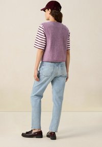 Femme portant un gilet tricoté violet sur une chemise rayée, un jean bleu clair, des mocassins bordeaux et une casquette bordeaux assortie, se tenant de dos.