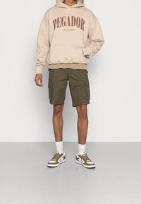 Felpa beige con testo "PEGADOR", indossata sopra una camicia chiara, abbinata a pantaloni cargo oliva e sneakers con dettagli marroni. Calzini bianchi visibili.