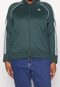 Mörkgrön zip-up jacka med vita ränder längs axlarna och ärmarna, två sidofickor, ribbade manschetter och krage.