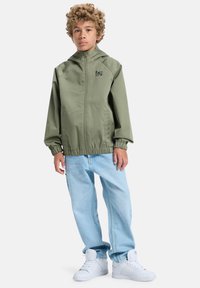 Oliivivihreä tuulitakki, jossa on vetoketju ja joustavat hihansuut, yhdistettynä vaaleansinisiiin farkkuihin ja valkoisiin tennareihin. Yksinkertainen logotyyli rinnassa.