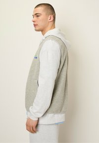 Light gray sleeveless vest with fleece texture, paired with a long-sleeve gray hoodie. A small colorful patch is visible on the vest.