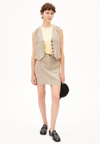 Beige geruite vest met ronde knopen over een geel shirt, gecombineerd met een bijpassende geruite rok. Zwarte instappers en een zwarte pet maken de look compleet.