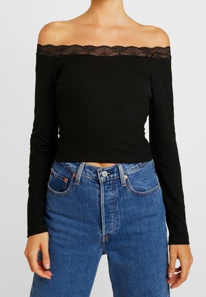 Femme portant un haut noir à manches longues épaules dénudées avec une bordure en dentelle et un jean en denim bleu taille haute, sur un fond uni.