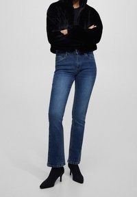 Person med mørkeblå slim-fit jeans, sorte sokkeboots og en sort fuzzy zip-up jakke med armene korslagt.