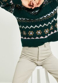 Henkilö, joka käyttää tummansinistä kuviollista villapaitaa ja vaaleita housuja, istuu valkoisella jakkaralla kädet ristissä pään yläpuolella.