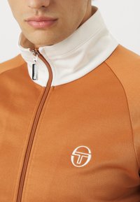 Oranžni zip-up jopič z belim ovratnikom, gladka tekstura, logo na prsih in kovinski podaljšek zadrge.