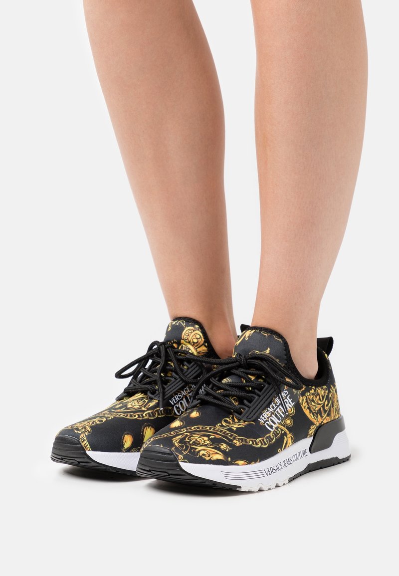 Svarta sneakers med ett guld barockmönster, rund tå, snördesign, vadderad sula och varumärke på sidan.