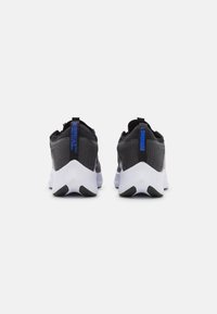 Paire de chaussures de course noires et gris foncé avec semelles blanches, vues de dos, montrant des languettes bleues avec texte sur fond blanc.