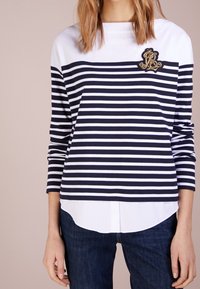 Donna che indossa una maglietta a righe orizzontali blu navy e bianca a maniche lunghe, con orlo bianco a strati e un ricamo dorato sul petto.