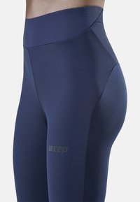 Marineblaue Hochbund-Leggings aus glattem, dehnbarem Material, mit einem Logo auf dem Oberschenkel und einem schlanken Design ohne sichtbare Nähte.