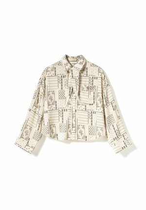 Beige pitkähihaisen paidan päällä mustia geometrisia ja kukkakuvioisia tilkkutöitä, etutasku ja solmittava kaulussomiste.