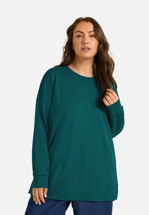 Kvinde med langt brunt hår iført en teal langærmet pullover og mørkeblå bukser, stående mod en lys baggrund.