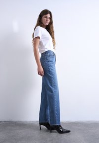 Νεαρή γυναίκα με μακριά μαλλιά, ντυμένη με λευκό t-shirt, μπλε τζιν με ψηλή μέση και μαύρες μυτερές μποτίνια με τακούνι, στέκεται μπροστά σε λευκό τοίχο.