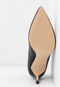 Chaussure noire à talon haut avec un bout pointu, tige en cuir lisse et une semelle en caoutchouc beige présentant des rainures horizontales discrètes. Agrémentée d'un logo.