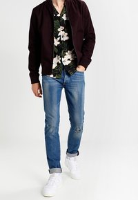 Man in versleten blauwe jeans, witte sneakers, een zwarte bloemenhemd en een donkere zip-up jas, met één hand in zijn zak, loopt vooruit.