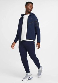 Marine atletisk tracksuit med lynlåslommer, glat overflade og tætsiddende design. Kombineret med sølvfarvede sneakers med blå detaljer.