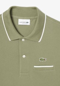 Polo verde oliva in tessuto texturizzato, con colletto bianco, due bottoni e un piccolo logo con coccodrillo ricamato sul taschino.