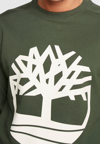 Felpa verde con colletto rotondo caratterizzata da un grande design di logo bianco di un albero, con una texture liscia e un colletto a coste per garantire durata.