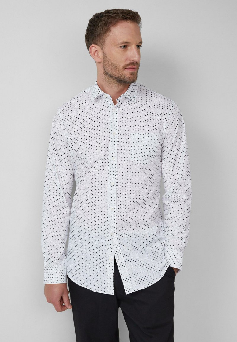 Homme portant une chemise blanche à manches longues avec de petits points noirs, un pantalon noir, se tenant devant un fond gris clair uni.