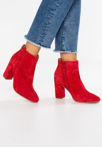 Bottines en daim rouge avec un talon carré, un bout pointu et une fermeture éclair. Un jean à ourlets effilochés met en valeur les chaussures, soulignant leur design.