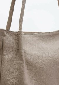 Beżowa skórzana torba tote z dwoma długimi paskami, o gładkiej fakturze i bezszwowej konstrukcji, z minimalistycznym wzornictwem i bez widocznych elementów metalowych.