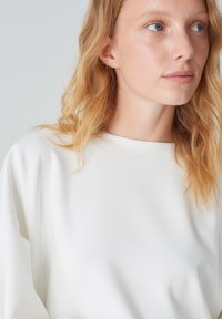 Maglia bianca a maniche lunghe con scollo rotondo e vestibilità ampia, realizzata in tessuto morbido e caratterizzata da cuciture sottili lungo i bordi.