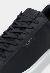 Nubikk sneakers met een marineblauwe leren bovenkant, gestructureerde veters, een subtiele gouden logo en een witte rubberen zool voor contrast.