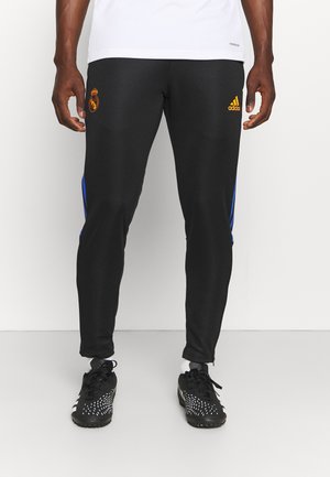Mann trägt schwarze Adidas Real Madrid Trainingshose und schwarze Fußballschuhe, steht vor weißem Hintergrund.