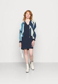 Vestido azul marinho com decote em V, acompanhado de uma jaqueta bomber multicolorida com padrões geométricos. Botas de tornozelo brancas com detalhes pretos.