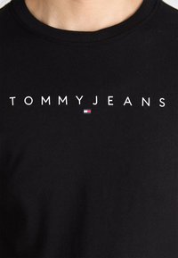 Crna pamučna majica s natpisom "TOMMYJEANS" u bijeloj boji, s malim naglaskom u crvenoj, bijeloj i plavoj boji ispod natpisa.