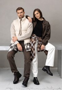 Dwie modelki mają na sobie warstwowe stroje: kremową pikowaną kamizelkę, brązową skórzaną kurtkę, koszulę flanelową w kratę, ciemnobrązowe dżinsy i czarne buty.